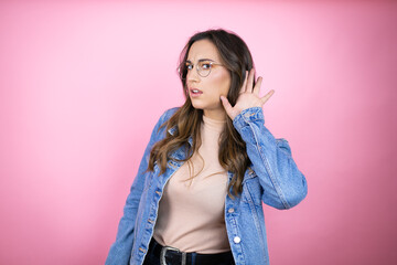 Young beautiful woman wearing denim jacket over isolated pink background surprised with hand over ear listening an hearing to rumor or gossip
