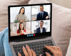 A woman is participating in a video conference meeting on a laptop. She is sitting on a couch and typing on the keyboard while looking at the screen, cropped