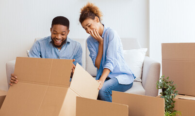 Relocation. Happy Afro Spouses Unpacking Moving Boxes Sitting On Couch In New House. Selective Focus