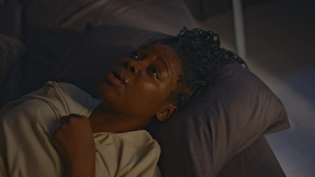 High angle shot of young Black woman lying sleepless in bed at night and calming herself down with slow breathing after having dreadful nightmares about catastrophic past events