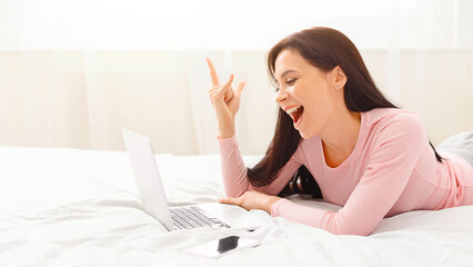 A delighted woman lies on her stomach on a white bed, with a laptop in front of her. She appears ecstatic, making a victory gesture with her hand, possibly after receiving good news