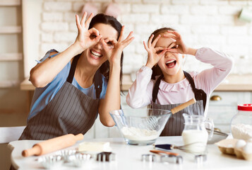 A happy mother and daughter are making cookies in the kitchen. They are both wearing aprons and are smiling as they look at the camera. They are making a mess of flour, but they are having fun.