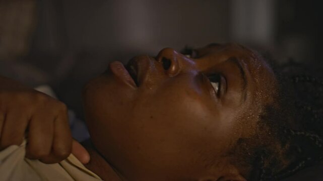 Side closeup of African American woman with trauma panting after having awful nightmare, lying in bed with sweat on forehead at night