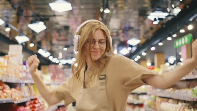 An enthusiastic employee, wearing a supermarket uniform, is joyfully dancing in the aisle with headphones on. They seem to be enjoying their work and showing positive energy