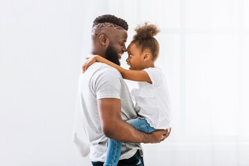 Fototapeta premium A Black father, with a beard, is smiling and holding his young daughter in his arms. The girl is also smiling and has her arms wrapped around her fathers neck.