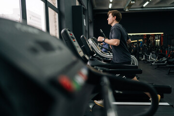 Side view of beginner sportsman having intensive cardio workout to burn calories in gym. Side view of young athletic man jogging on gym machine, having cardio session in well-equipped fitness club.