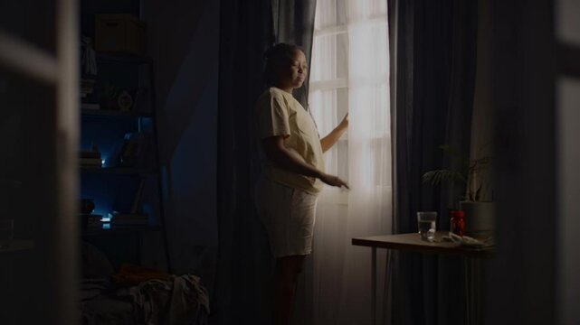 Full shot of young Black woman looking out of window at dawn and walking around room on sleepless night. Avoiding real life, feeling lonely, living with ptsd