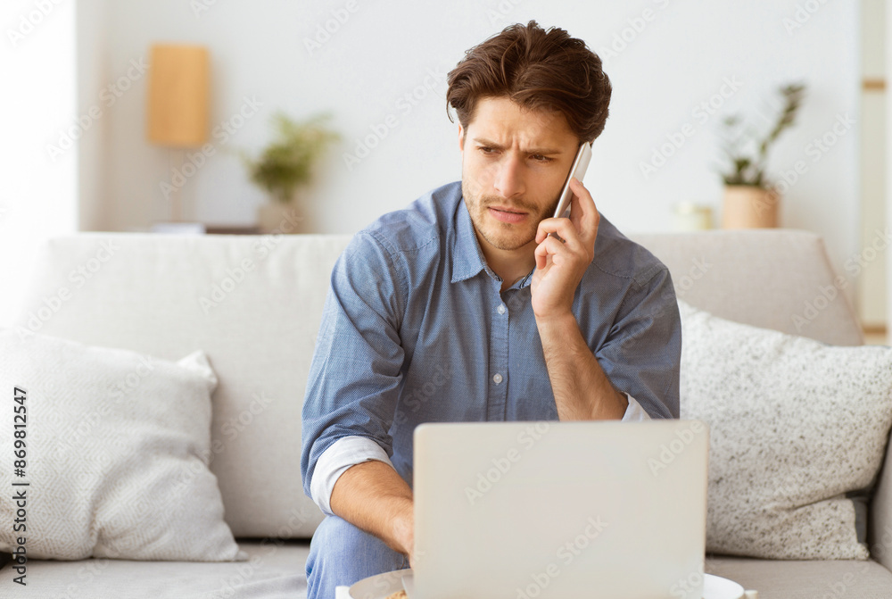 Poster a young man sits on a couch, working on his laptop while talking on his phone. he appears to be focu