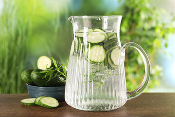 Refreshing cucumber water with rosemary in jug and vegetable on wooden table against blurred green...