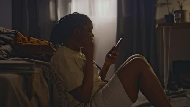 Side shot of young stressed Black woman sitting on floor by bed and getting irritated while talking on phone at home. She throwing mobile away and feeling tense