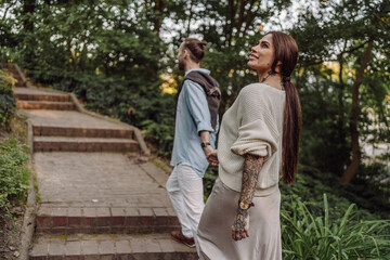 Fototapeta premium Outdoor shot of young couple in love walking on pathway through grass field. Young couple strolling in the park. Trendy couple with tattoos walking in a park. Botanic Garden.