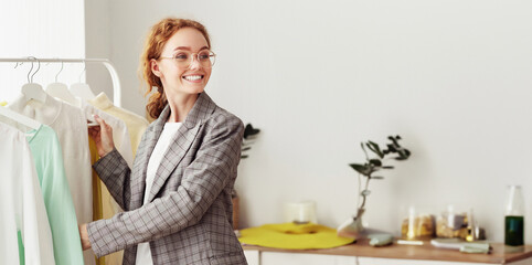 Fashion Closet Concept. Happy stylish girl in glasses choosing designer clothing on hangers, copy space