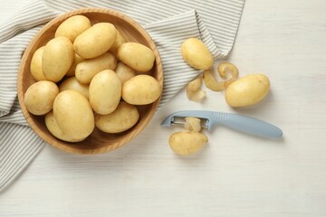 Fresh raw potatoes, peels and peeler on white wooden table, flat lay