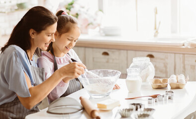 A mother and daughter are baking together in a bright kitchen. The daughter is stirring a bowl of flour with a whisk, while the mother watches with a smile. They are both wearing aprons