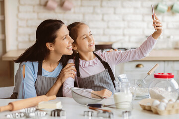 A mother and her young daughter are taking a selfie in the kitchen. They are both wearing aprons and smiling. There are baking supplies on the table in front of them