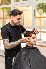 tattooed barber cutting the hair and beard of a young, dark-haired client.
