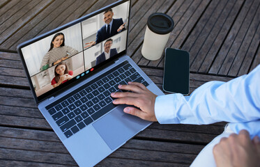 A person is sitting outdoors at a table and using a laptop to participate in a video conference call. The laptop screen shows multiple people on a video call.