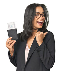 Young Latina in business suit with passport points with thumb finger away, laughing and carefree.