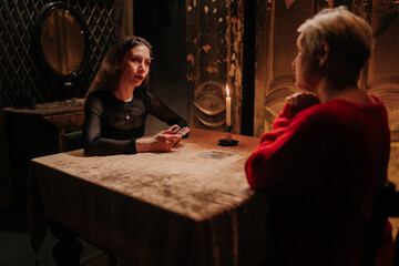 Rear view of fortuneteller doing Tarot card reading and talking to unhappy adult woman client about...