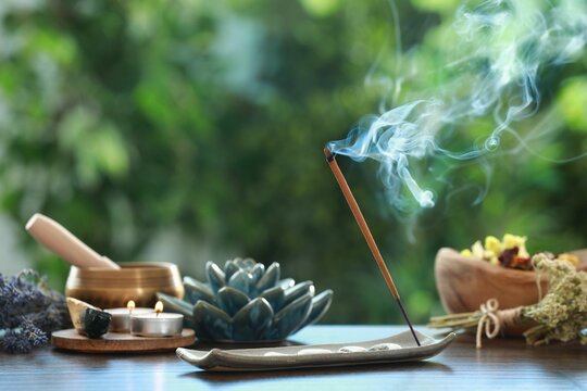 Incense stick smoldering in holder, burning candles, dry flowers and Tibetan singing bowl on wooden table outdoors