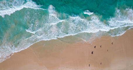 Aerial view of a sandy beach with turquoise waves crashing onto the shore scattered people are seen walking along the coastline