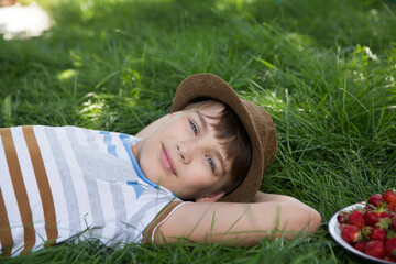 Happy  preteen boy laying on grass with a strawberry basket in summer. Happy ghild having fun outdoors. Generation Z. Happy childhood.
