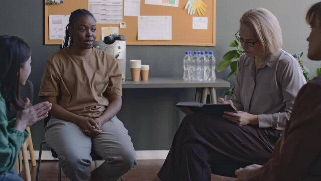 Medium long pan shot of female African American war veteran sharing traumatic memories from her military service during mental health support group meeting, Caucasian therapist listening attentively