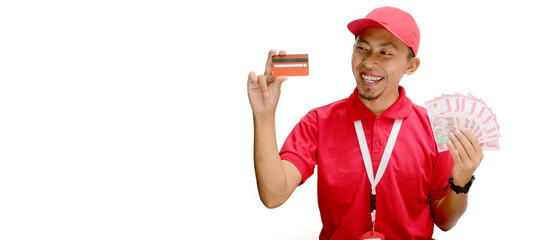 Asian delivery man holds a credit card and Indonesian Rupiah banknotes against a white background. Delivery services, financial transactions, e-commerce, and customer satisfaction.