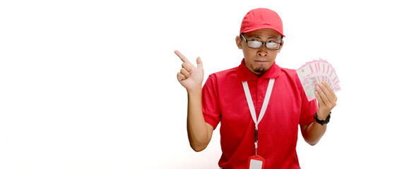 Excited Asian delivery man or courier points excitedly at empty copy space for text or advertisement, while holding money banknotes against a white background.