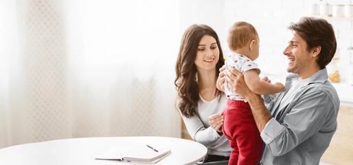 A young family is enjoying their time together in the kitchen. The father is holding their baby in his arms, while the mother smiles at them. Baby is looking up at his father with a smile, copy space
