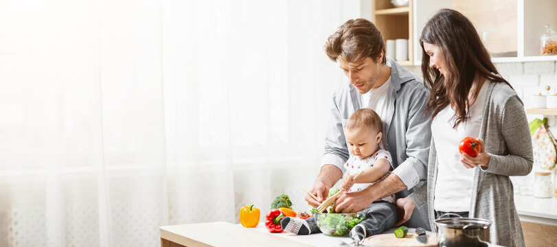 A family of three is preparing food together in a kitchen. The father is holding their baby while they all help prepare vegetables. The mother is standing next to them, copy space