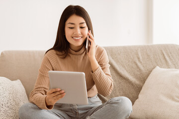 A woman sits on a couch, using a tablet and talking on the phone.