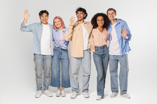 Five young adults, including three men and two women, stand together in a studio setting, smiling and waving to the camera. They are all wearing casual clothes and appear to be happy