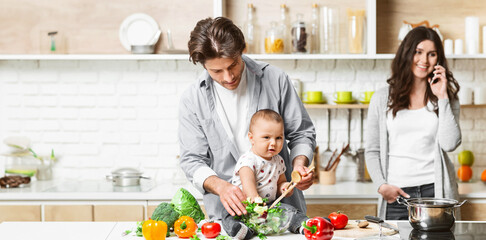 A father cooks in the kitchen while holding his baby, who is sitting on the counter. The mother is standing behind the counter, talking on the phone, copy space