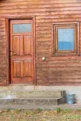 Forest Cabin House. Entrance brown  Door and Window Frame with Wooden Planks Background and Plastic Garbage Bin by Concrete Stairs.