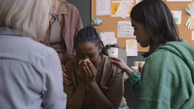 Medium shot of psychologist and members of ptsd support group comforting young woman who crying after sharing her traumatic memories, therapist giving napkin to patient
