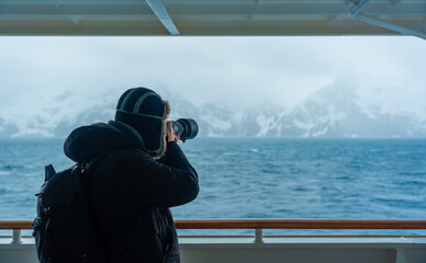 Male Photographer on Antarctica Cruise Ship Expedition Professional Telephoto Lens Railing View of Icebergs Mountains Warm Hat Backpack Black Coat © And They Travel