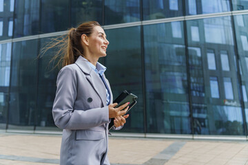 A businesswoman in a grey suit holds a phone and a tablet, smiling while standing outside a modern...