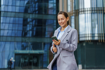 A businesswoman in a grey suit smiles while using her phone, standing in front of modern glass buildings.