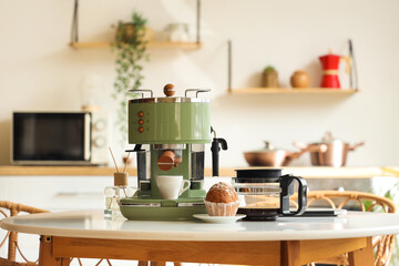 Modern coffee machine with cup, cupcake and pot on table in kitchen
