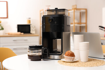 Modern coffee machine, muffin, paper cups and laptop on white table in kitchen