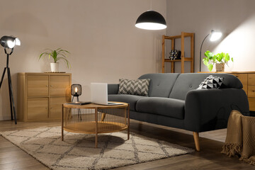 Interior of living room with comfortable sofa, table, modern laptop and glowing lamps