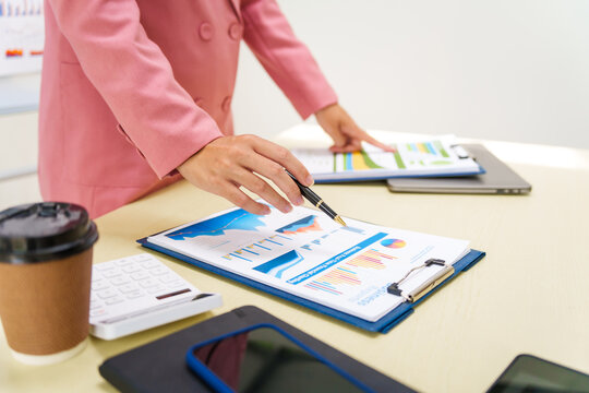 A businesswoman in a pink suit works at a desk with a whiteboard in the background. She focuses on accounting and financial services, professionalism and expertise in various financial roles. - Powered by Adobe