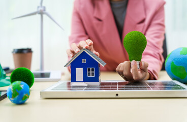 A businesswoman in a pink suit stands at a white desk with a world model, electric car model, and green light bulb, promoting Earth Day, green energy, and sustainability efforts.