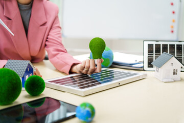 A businesswoman in a pink suit stands at a white desk with a world model, electric car model, and green light bulb, promoting Earth Day, green energy, and sustainability efforts.
