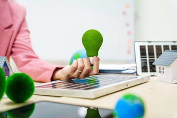 A businesswoman in a pink suit stands at a white desk with a world model, electric car model, and green light bulb, promoting Earth Day, green energy, and sustainability efforts.