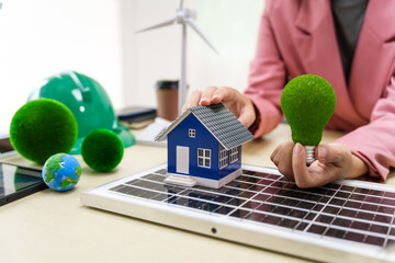 A businesswoman in a pink suit stands at a white desk with a world model, electric car model, and green light bulb, promoting Earth Day, green energy, and sustainability efforts.