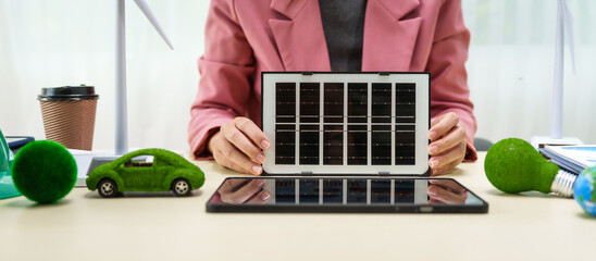 A businesswoman in a pink suit stands at a white desk with a world model, electric car model, and green light bulb, promoting Earth Day, green energy, and sustainability efforts.