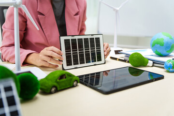 A businesswoman in a pink suit stands at a white desk with a world model, electric car model, and green light bulb, promoting Earth Day, green energy, and sustainability efforts.