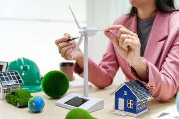 A businesswoman in a pink suit stands at a white desk with a world model, electric car model, and green light bulb, promoting Earth Day, green energy, and sustainability efforts.